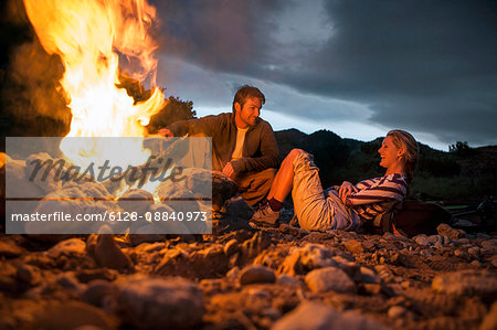 Young couple relaxing and laughing beside glowing campfire.