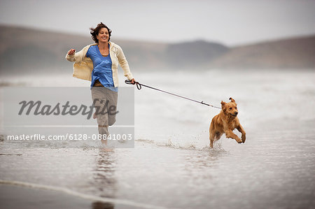 Smiling mid adult woman running along a beach with her dog.