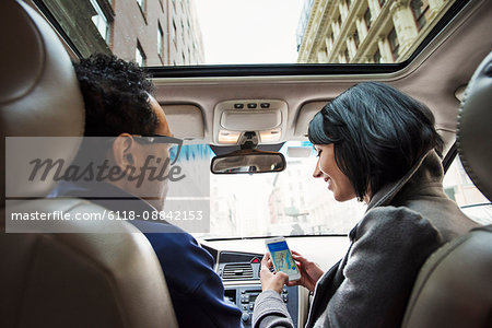 A young woman and young man in a car looking at a map on the display of a cellphone, seen from the back seat.