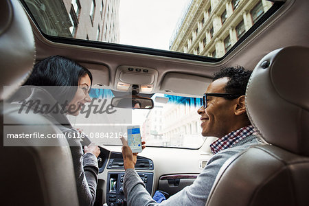 A young woman and young man in a car looking at a map on the display of a cellphone, seen from the back seat.