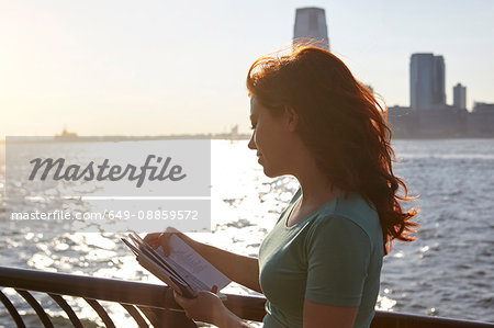 Young female tourist with long red hair looking at guidebook on waterfront, Manhattan, New York, USA