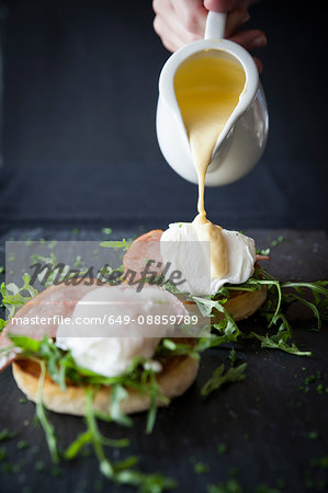 Female hand pouring hollandaise sauce over eggs benedict breakfast on slate