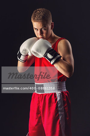 Young handsome boxer sportsman in red boxer suit and white gloves standing on black backgound. Copy space.