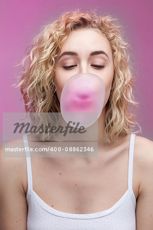 studio closeup portrait of blond woman with curly hair doing a bubble with a chewing gum on a pink background