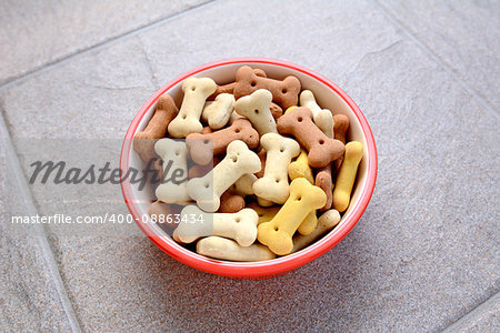 Red pet food bowl full of bone-shaped dog biscuits, on a grey tile floor