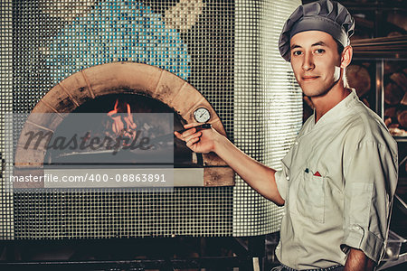 Young Italian chef in white uniform and hat in the kitchen interior. Man holding a shovel with a raw pizza and going to put it in a special oven.