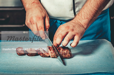 Chef in hotel or restaurant kitchen dressed in blue apron cooking, only hands, he is cutting meat or steak with big sharp knife