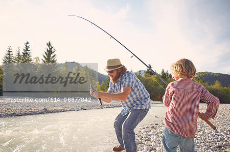 Mid adult man and boy next to river using fishing rod to catch fish