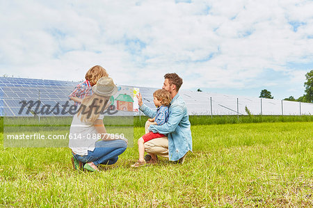 Young family sitting in field, looking at child's drawing of house, next to solar farm