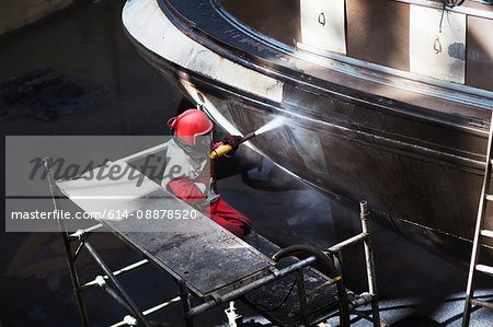 Worker cleaning hull of boat with high pressure hose in shipyard