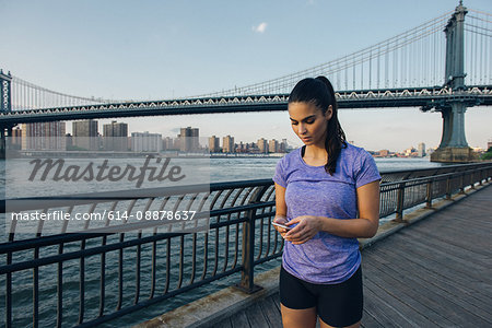 Young female runner reading smartphone in front of Manhattan bridge, New York, USA