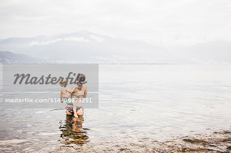 Rear view of mother standing in lake holding boy looking away at view of mountain range, Luino, Lombardy, Italy