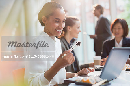 Smiling businesswoman eating sushi lunch with chopstick at laptop in conference room meeting