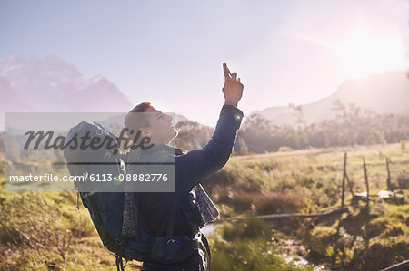 Young man with backpack hiking and using camera phone in sunny, remote field