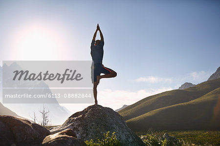 Young man balancing in tree pose on rock in sunny, remote valley