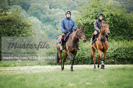 Two men on bay horses riding across a field side by side. Woodland.