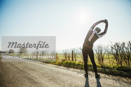 A man preparing for a cycle ride, with raised arms stretching and leaning sideways. A country road.