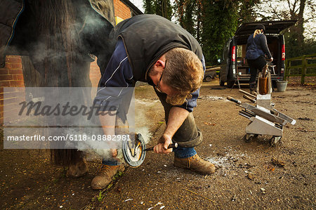 A farrier shoeing a horse, bending down and fitting a new horseshoe to a horse's hoof.