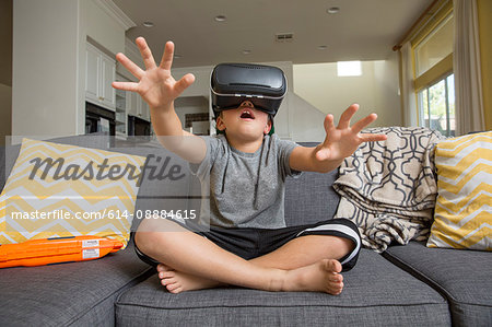 Young boy sitting cross legged on sofa, wearing virtual reality headset, hands reaching out in front of him