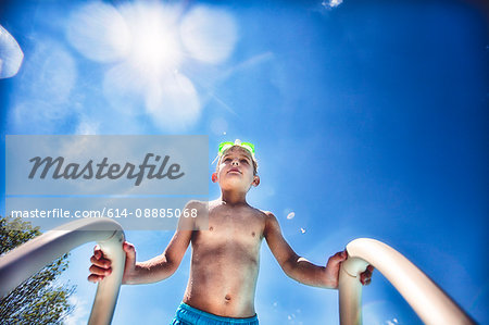 Low angle view of boy on swimming pool ladder against blue sky