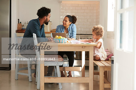 Girl Paints At Kitchen Table As Parents Look At Laptop