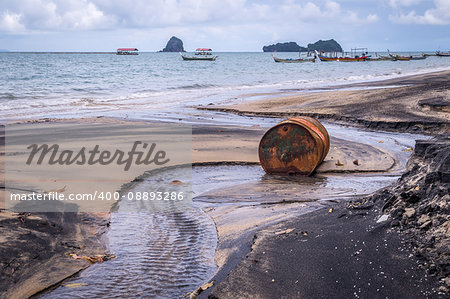 Rusty barrel oil on a partly black coloured beach illustrates the pollution of environment by oil spills. Longtail boats in the background.