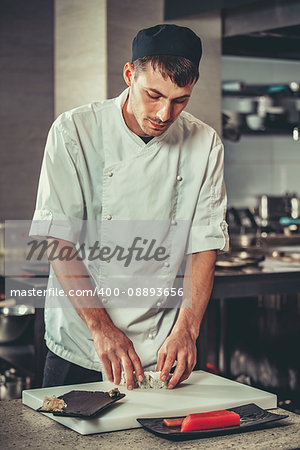 Food: One young white chef dressed in white uniform decorate ready dish in restaurant. He is working on maki rolls. Preparing traditional japanese sushi set in interior of modern professional kitchen