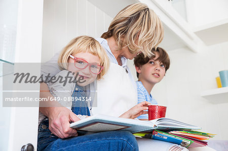 Mature woman sitting on kitchen counter reading storybooks with son and daughter