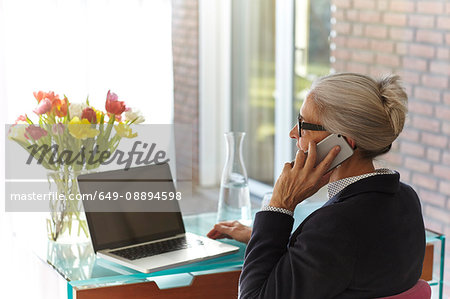 Senior businesswoman using laptop and making smartphone call at home desk