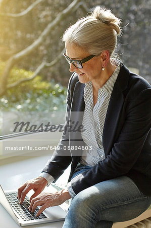 Senior businesswoman typing on laptop at home on windowsill