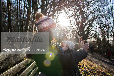 Mother sitting on bench with baby boy, taking selfie with smartphone