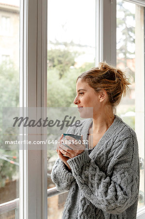 Woman at home, looking out of window, holding hot drink