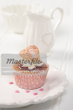 A sweet heart cupcake on a white plate with white milk jug and sugar basin in background