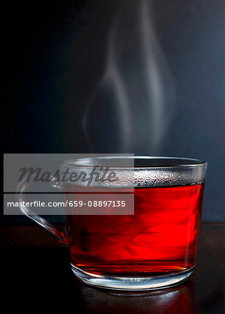 Hot red berry fruit tea in a glass mug with steam standing on a dark wood with dark grey background