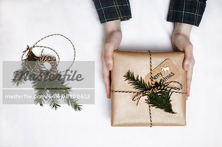 Woman holding Christmas gift wrapped in brown paper, decorated with fern and string, overhead view