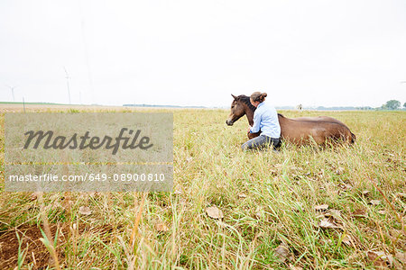 Woman crouching with arm around horse lying down in field