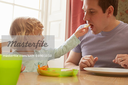 Female toddler feeding bread to father at kitchen table