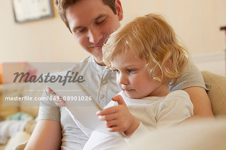 Female toddler sitting on sofa with father using digital tablet