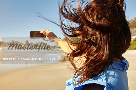 Mature woman on beach, taking selfie, using smartphone, rear view