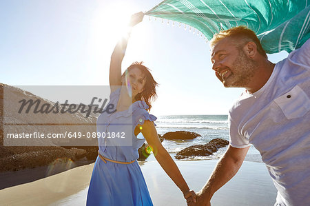 Mature couple holding hands on beach, woman holding sheer scarf in air