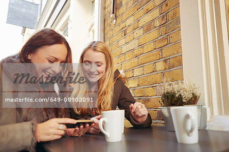 Two female friends, sitting outdoors, having coffee, looking at smartphone