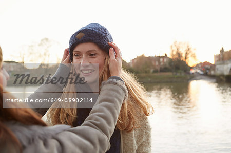 Young woman putting knitted hat on friend, outdoors, smiling