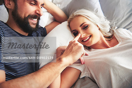 Couple lying in bed, fooling around, man poking woman's nose