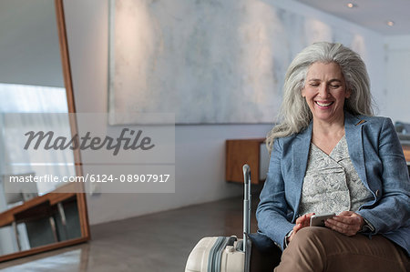 Smiling mature woman with suitcase texting with cell phone