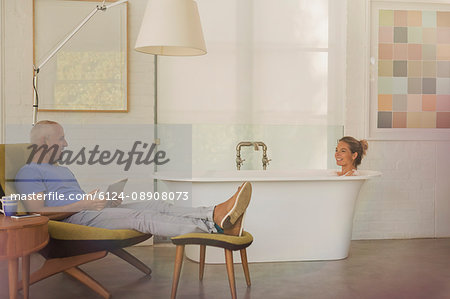 Husband with digital tablet relaxing, talking to wife in soaking tub in luxury hotel room