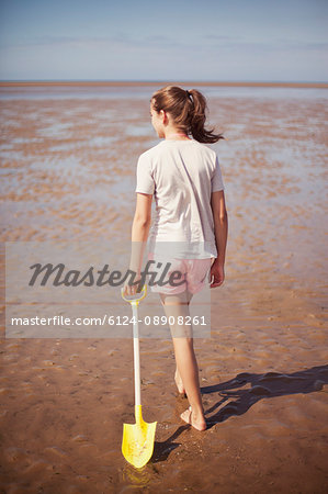 Teenage girl dragging shovel in wet sand on sunny summer beach
