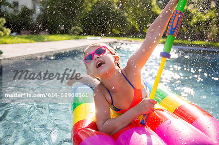 Girl in bikini on inflatable playing with water gun in outdoor swimming pool