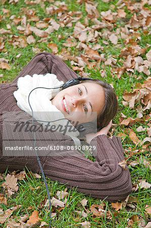 Portrait of a content woman lying down in the park smiling at camera and listening to music