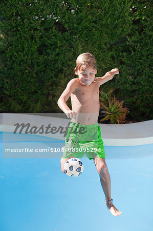 Young boy jumping into swimming pool kicking ball