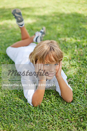 Cute blonde boy lying down on grass in park smiling at camera